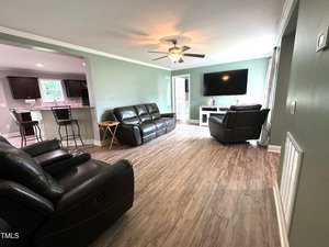 Spacious living room with light green walls, featuring a ceiling fan and wooden flooring. It includes two leather sofas, a recliner, a large flat-screen TV on a white stand, and a view into the kitchen area with bar stools.