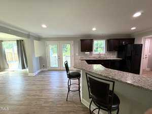 A modern kitchen with granite countertops, a curved breakfast bar, and two barstools. The kitchen has dark cabinets, a black refrigerator, and wooden flooring. Large glass doors lead to an outdoor area with natural light streaming in.