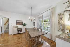 A bright dining room with a wooden table and six chairs, a chandelier overhead, large window with curtains, and a sideboard holding books and a plant. A staircase is visible in the background. White walls and wood flooring complete the space.