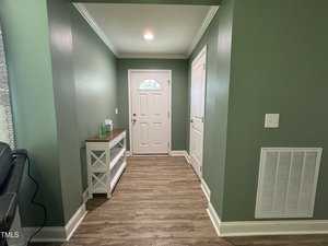 A hallway with green walls and wood flooring. It features a white front door with a small half-circle window, a white utility door, and a white console table with a sanitizer bottle and towels on it. Natural light from a window on the left.