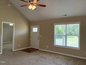 A room with beige walls and carpet, featuring a ceiling fan with lights. There is a white door and a large window on one wall, allowing natural light in. An adjoining room is visible through an open doorway.
