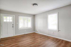 A bright, empty room with light gray walls and two windows with blinds. The floor is made of wood planks, and a white front door is in the corner. A ceiling light fixture is centered above.