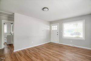 Empty room with light gray walls, wood laminate flooring, and a window with blinds. A white door with glass panels is on the right, and a small bathroom is partially visible through a doorway on the left. Ceiling light fixture is centered.