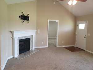 A living room with beige walls features a white fireplace, an empty TV mount above it, and a ceiling fan. There's a doorway leading to another room and a door with a window nearby. The carpet is light brown.