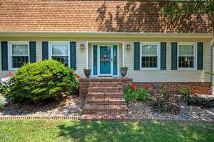 A charming house with a brown shingle roof, light-colored siding, and dark shutters. Three steps lead to a teal front door adorned with a wreath. The yard features green shrubs and blooming red flowers.