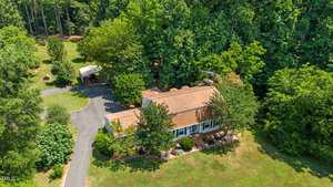Aerial view of a single-story house with a brown roof, surrounded by lush greenery and tall trees. A paved driveway leads to a carport on the left. The yard is well-maintained with shrubs and grassy areas.
