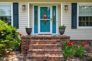 A house entrance with a white door featuring decorative glass and a flower wreath. The steps are made of red brick, flanked by potted plants and red flowers. The exterior wall is light-colored with two windows adorned with dark shutters.