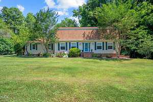 The charming single-story house at 201 Holly Drive in Oxford features a brown roof and beige siding, nestled among lush green trees and lawn. A brick walkway leads to the welcoming front door adorned with a wreath, while flowers beautifully line the windows.