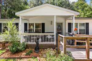 A white mobile home features a covered porch with white railings and a ramp leading up to the entrance. There's a statue of a dog in the garden on the left, surrounded by greenery and mulch. Trees can be seen in the background.