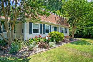 A charming single-story house with beige siding and dark shutters, surrounded by lush landscaping. Red flowers and a variety of plants are in the garden bed. A pathway of brick steps leads to the entrance. Large trees provide shade in the background.