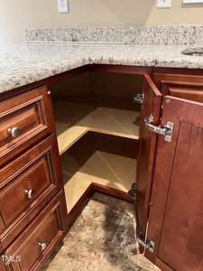 Open kitchen cabinet with brown wood finish and three visible shelves. Adjacent countertop is light-colored granite. Surrounding flooring is brown tile. Cabinet doors are open, revealing empty storage space.