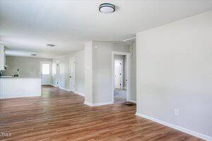 A modern, unfurnished open-concept living space with light wood flooring and light gray walls. The room features recessed lighting, a view into a white kitchen with an island, and several doors leading to other areas. Natural light fills the space.