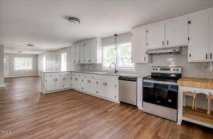 Spacious kitchen with white cabinets, stainless steel appliances, and wood flooring. Large windows provide natural light. Open layout extends to dining and living area.