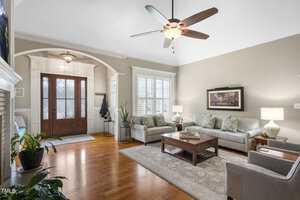 Spacious living room with hardwood floors, light gray sofas, and armchairs. A ceiling fan hangs above. There's a wooden coffee table, a large painting on the wall, and plants by the entrance with a coat rack and decorative light fixture.