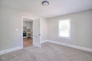 Empty room with light gray walls and textured carpet. A single window lets in natural light. An open door reveals a glimpse of a kitchen area with wooden flooring, white cabinets, and an oven. A simple ceiling light fixture is installed.