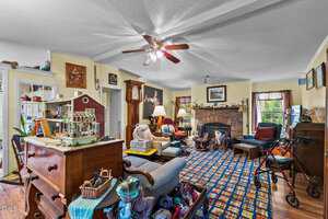 A cluttered living room filled with various items, including a wooden chest, model house, toys, and a walker. The room has a patterned carpet, white ceiling fan, fireplace, and a mix of eclectic decor on the walls. Chairs and tables are scattered around.
