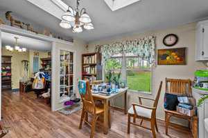 A cozy dining area with wooden floor and furniture. The room is decorated with floral curtains and various decorations on the shelves. A table and chairs are set by a window showcasing a view of a green yard outside. Natural sunlight fills the space.