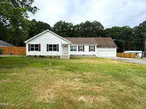 A white single-story house with a front porch and attached two-car garage. The yard is expansive and grassy, with a few trees in the background. A wooden fence lines part of the property.