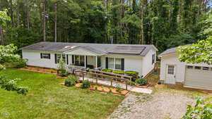 A white single-story house with a ramp leading to the front porch, surrounded by trees and greenery. A detached white garage is on the right, with a gravel driveway extending from it. Solar panels are visible on the house roof.