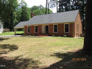 A brick house with a dark gray roof and chimney, surrounded by a grassy yard with tall trees in the background. The house has a small porch with steps leading up to the front door. The photo is dated June 20, 2024.