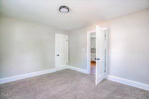 A bright, empty room with light gray walls and carpeted flooring. There are two closed white doors and a ceiling light fixture. Sunlight is streaming in through an unseen window, casting a soft illumination.