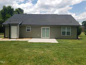 A small, single-story house with a dark roof and light-colored siding. The house has a back door with a small patio and a grassy backyard. The sky is partly cloudy.