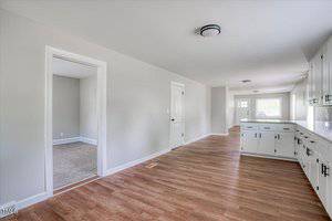 A spacious, empty hallway with hardwood floors, light gray walls, and ceiling lights. On the left, there's a doorway leading to a carpeted room. On the right, white cabinets with a countertop line the wall. Natural light filters in from windows at the end.