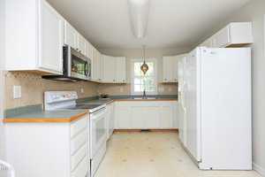 A bright, narrow kitchen with white cabinets and appliances, including a refrigerator and stove. There is a small window above the sink, and light-colored tiles on the floor and backsplash.