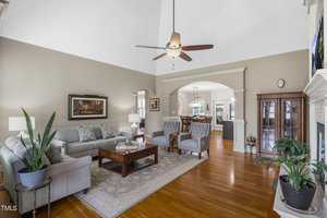 Bright living room with vaulted ceiling, chandelier fan, hardwood floors, and gray walls. Includes a gray sofa, two armchairs, coffee table, and potted plants. An archway leads to the dining area with a table and chairs in the background.