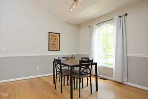 A minimalist dining room with a wooden floor features a square table and four chairs. A large arched window with white curtains allows natural light to fill the room. A framed artwork decorates the light gray walls with white trim.
