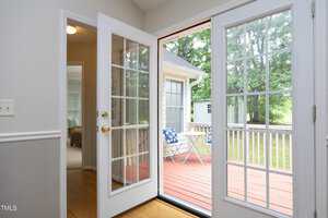 Open French doors leading to a wooden deck with a small table and chairs, overlooking a grassy backyard. White interior walls and wooden flooring inside, with a glimpse of a cozy room. Trees are visible in the background.