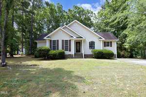 Nestled on 1992 Ferbow Street in Creedmoor, this charming single-story white house features brown shutters and a dark roof. Surrounded by lush green shrubs and trees, it boasts a small front porch and sits peacefully back from the grassy yard under a partly cloudy blue sky.