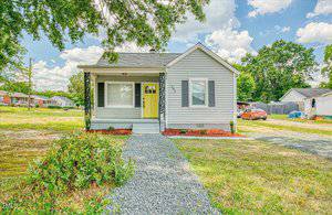 A cozy, single-story gray house with white trim and a bright yellow front door sits charmingly on 201 Harris Street, Oxford. The house features a black-shuttered window and a pathway leading to the entrance. Surrounded by a grassy yard, neighboring houses are visible in the background.