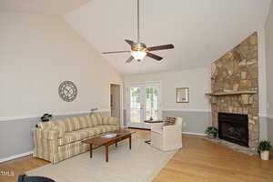 A cozy living room with a striped sofa, wooden coffee table, and a single chair arranged on a light rug. The room features a stone fireplace, a ceiling fan, and French doors leading to a patio. A decorative clock and mirror adorn the walls.