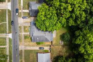 Aerial view of a suburban neighborhood featuring a street lined with several houses. The central house is surrounded by lush green trees. A small patio area is visible in the backyard along with a driveway and parked cars.