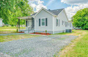 A small, single-story house with light gray siding and a dark roof is surrounded by a gravel driveway and green lawn. There are a few shrubs near the foundation. The sky is partly cloudy with sunlight illuminating the scene.