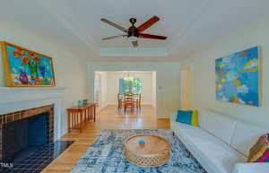 A cozy living room with a white sofa, colorful cushions, and a round wooden coffee table on a patterned rug. A ceiling fan hangs above. Bright paintings adorn the walls. A fireplace is on the left, and a dining area is visible through an open doorway.