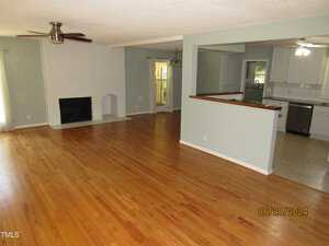 A spacious open-concept living area featuring hardwood floors and a white brick fireplace. The room leads into a kitchen with white cabinets, stainless steel appliances, and tiled flooring. A ceiling fan is mounted above the living space.
