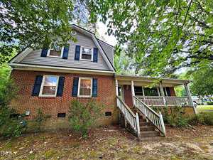 A charming two-story brick house with blue shutters and a gabled roof sits gracefully at 100 Royall Road. The front porch features white railings, with stairs leading up to the entrance, evoking classic Oxford elegance. Lush trees and greenery envelop this picturesque home.