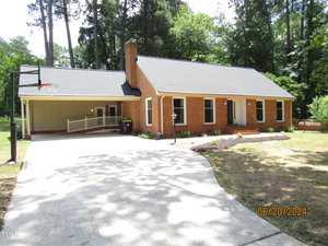 A red brick one-story house with a black roof, a chimney, and a partially covered driveway. A basketball hoop is installed on one side of the driveway. The house is surrounded by tall trees and a grassy yard. The date stamp reads 06/20/2024.