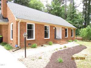 A brick house with a sloped roof, white-framed windows, and a central white door. There's a gravel path leading to the entrance, surrounded by a lawn and trees. A lamppost is near the walkway. Date-stamped as June 20, 2024.