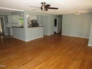 Spacious living area with wooden floors, light gray walls, and a ceiling fan. There's an open kitchen with white cabinets and stainless steel appliances. The room is well-lit, with large windows on the left, and a chandelier in the distance.