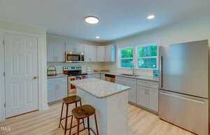 Modern kitchen with white cabinets, granite countertops, and a central island with two stools. Stainless steel appliances include a refrigerator, oven, microwave, and dishwasher. Hardwood floor and large window provide a bright, inviting atmosphere.
