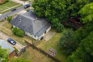 Aerial view of a residential property with a grey-roofed house, a backyard patio, and a fenced-in area. The backyard has seating around a fire pit and is surrounded by trees. A street and driveway are visible in the foreground.
