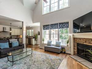 A spacious living room with a high ceiling, large windows, and a fireplace. The room features a sofa, glass coffee table, and TV. A modern kitchen with white cabinets is visible in the background. Natural light enhances the airy feel of the space.