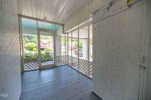 A bright, enclosed porch with white brick walls and a blue ceiling. The space features a wooden floor and lattice railing. Through the windows, a view of a garden with flowering bushes and a residential neighborhood can be seen.