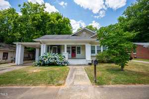 A single-story house with a covered porch and two white columns. The house is light gray with white trim and has a front yard with blooming hydrangeas and a small tree. A driveway is on the left, and the sky is partly cloudy.