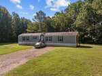 Located at 4752 Old Nc 75, this single-story mobile home in Oxford features a gray exterior and a quaint porch on a grassy lot. A gravel driveway guides you to a parked car, all framed by tall trees under a partly cloudy sky.