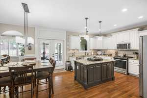 A modern kitchen with white cabinets and stainless steel appliances. An island with a granite countertop is in the center, surrounded by wooden chairs. The room is well-lit with pendant lights and has a dining area with a large wooden table.