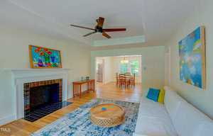 A living room with a white couch and a round wooden coffee table on a blue patterned rug. There's a ceiling fan above, colorful paintings on the walls, a fireplace, and an open view into a dining area with a table and chairs.